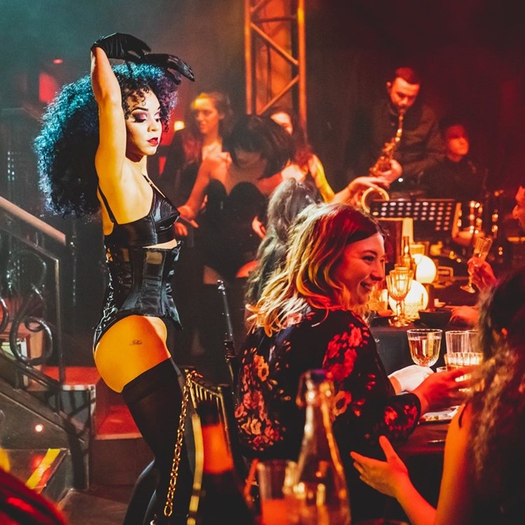 A black-corset cabaret performer with raised arms beside guests in warm red and gold light
